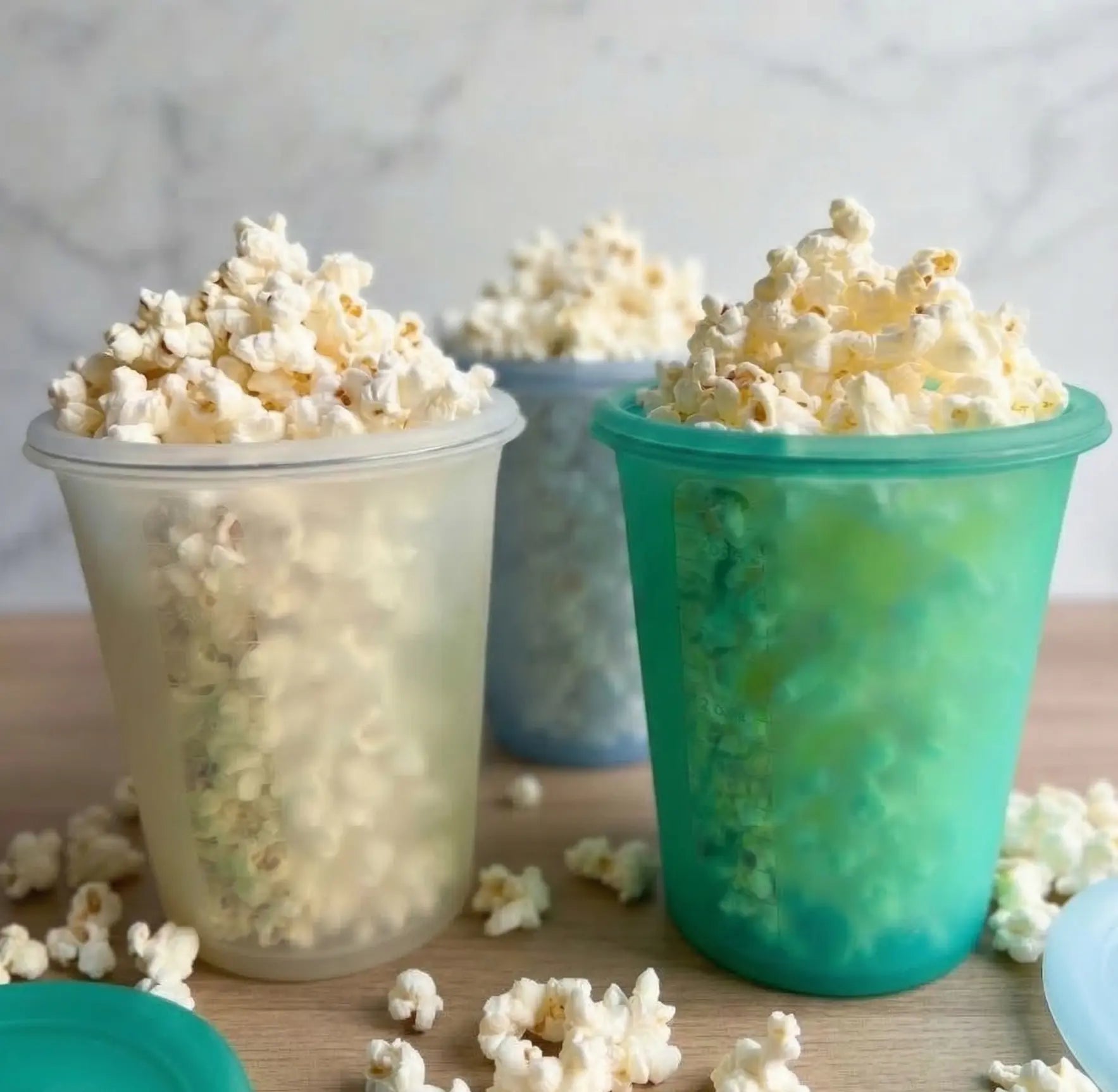 Three Anydelis filled with popcorn on a wooden surface in light blue, light basil, and opaque clear, with matching lids.