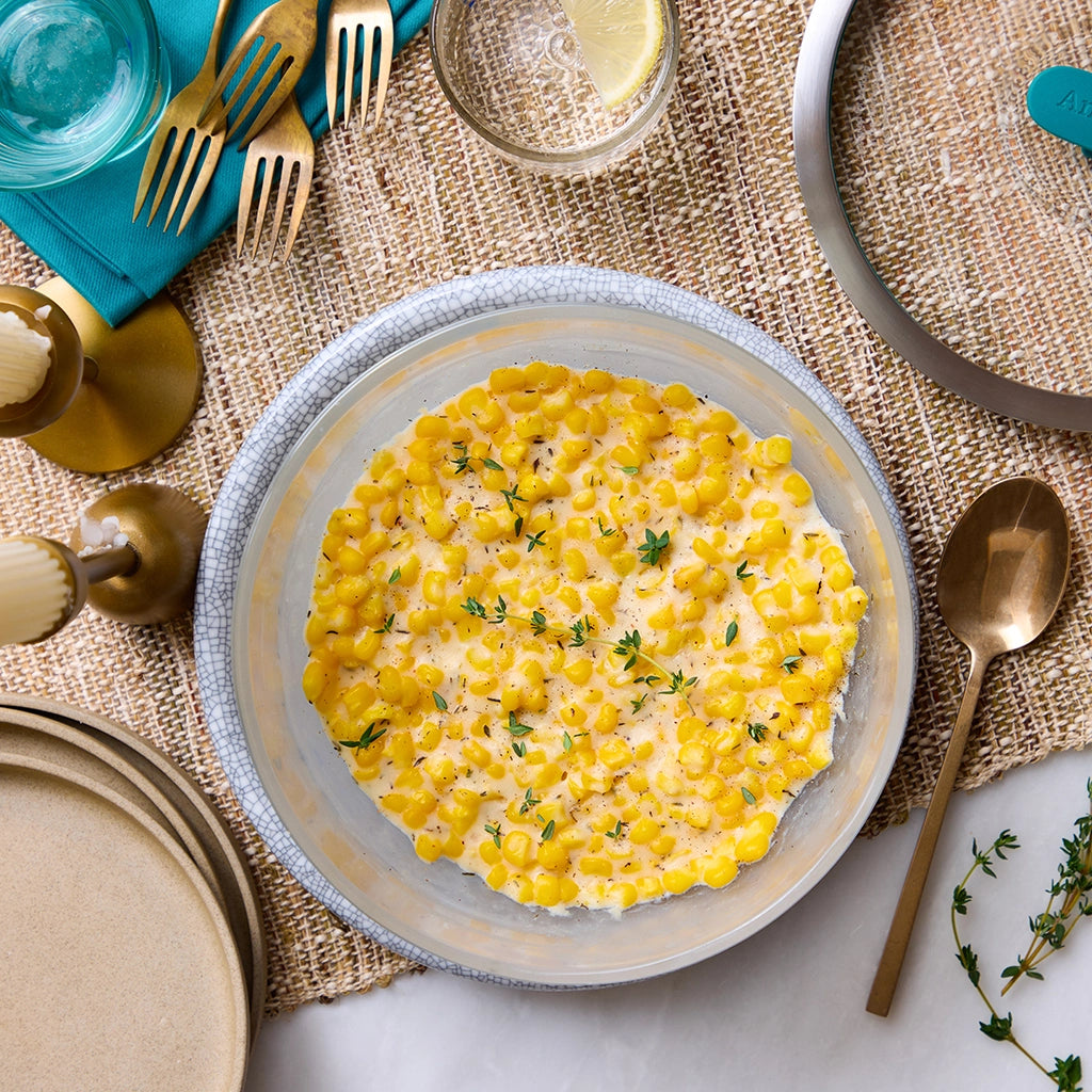 Brown butter creamed corn garnished with fresh thyme, made and served in a clear glass Anyday dish on a burlap table setting.