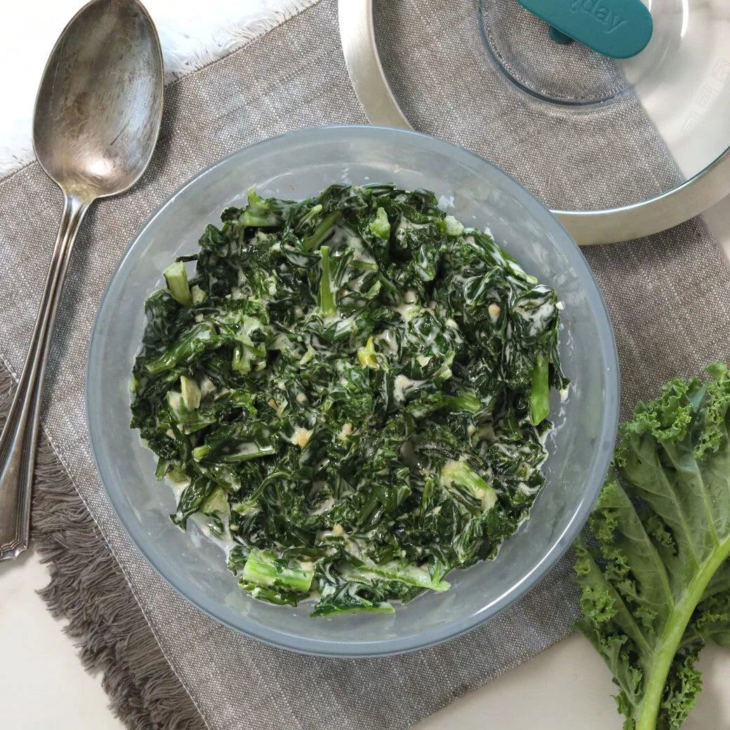 Creamed kale with garlic and seasoning served in a clear Anyday dish, placed on a linen napkin with a spoon and fresh kale leaves beside it.