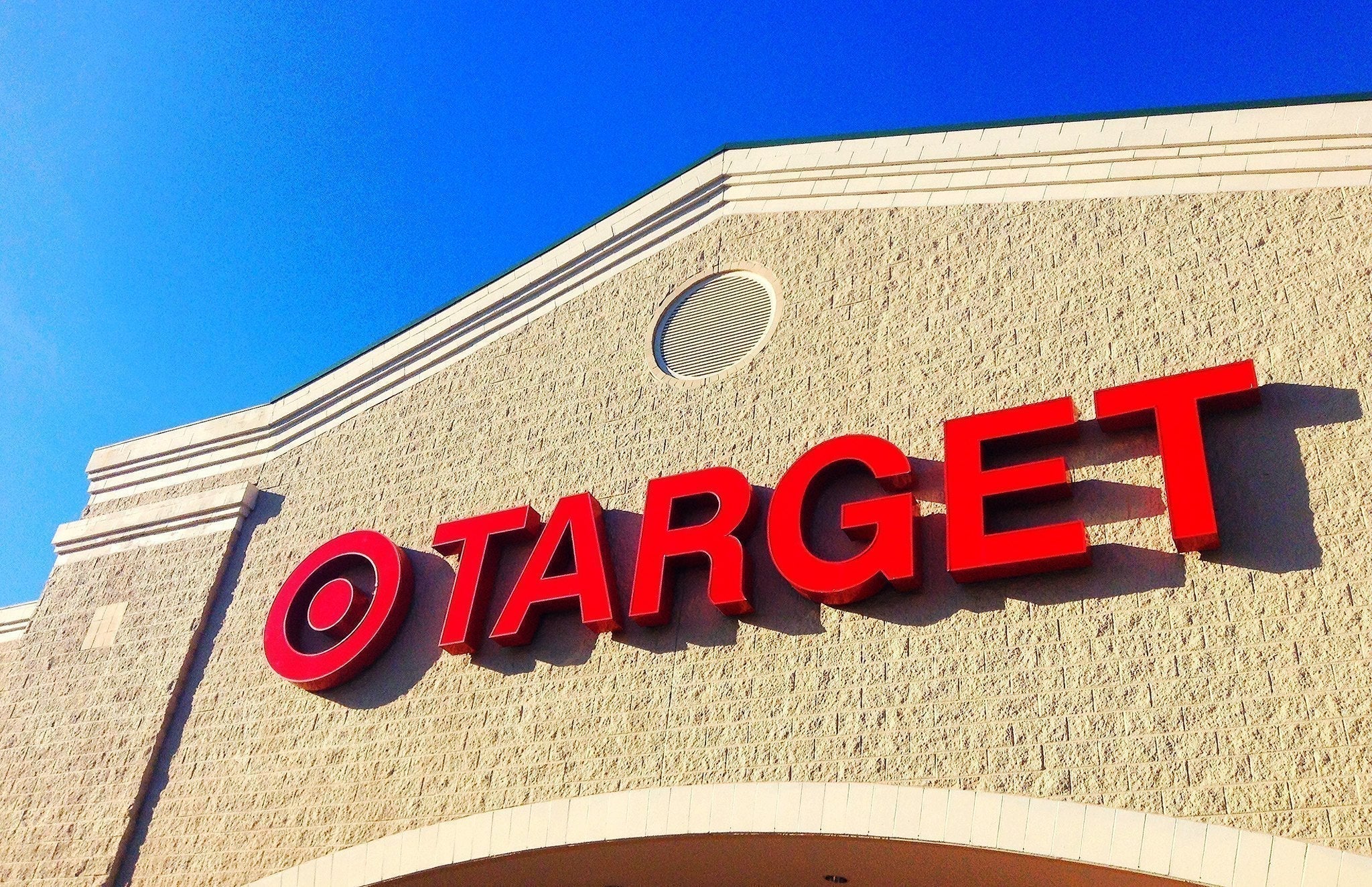 A Target store exterior with the red Target logo mounted on a beige brick wall. The bright blue sky contrasts with the bold red lettering.