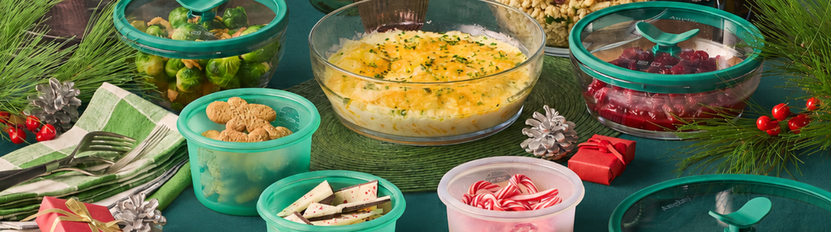 Glass food containers with teal lids hold Brussels sprouts, potato casserole, cranberries, gingerbread, chocolate bark, and peppermint candies on a green tablecloth with holiday greenery.