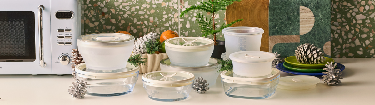 Assorted clear glass food storage containers with white lids arranged on a kitchen counter next to a light blue microwave and holiday decor.
