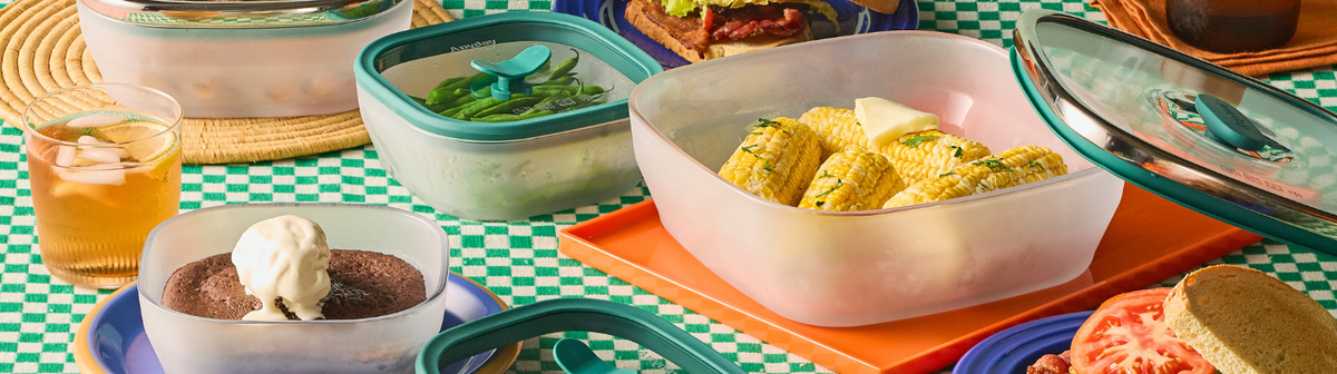 Plastic containers filled with green beans, corn on the cob, and chocolate cake with ice cream sit on a checkered table, alongside iced tea and a sandwich with tomato and lettuce.