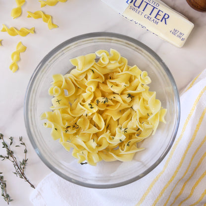 Microwave buttered egg noodles with fresh thyme in an Anyday glass dish, with a stick of butter on the counter and herbs nearby.