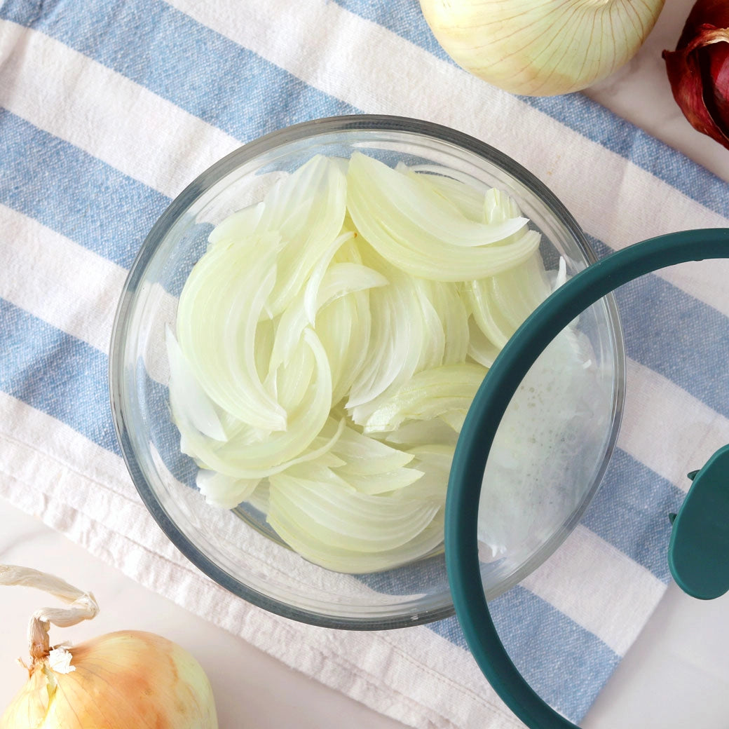 Lightly sautéed onions in an Anyday glass dish with lid, resting on a striped kitchen towel.