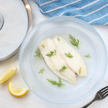 Cooked white tilapia fillet in a clear Anyday dish with herbs and lemon slices


