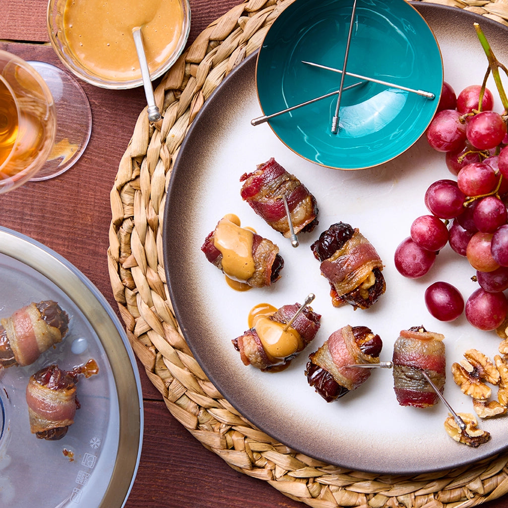 Bacon-wrapped walnut dates served on a plate with fresh grapes and a side of dipping sauce, prepared in a clear glass Anyday dish.