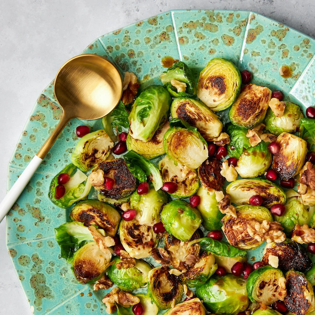 Balsamic charred Brussels sprouts garnished with pomegranate seeds and walnuts, make and in a glass Anyday dish and served on a green plate with a gold serving spoon.