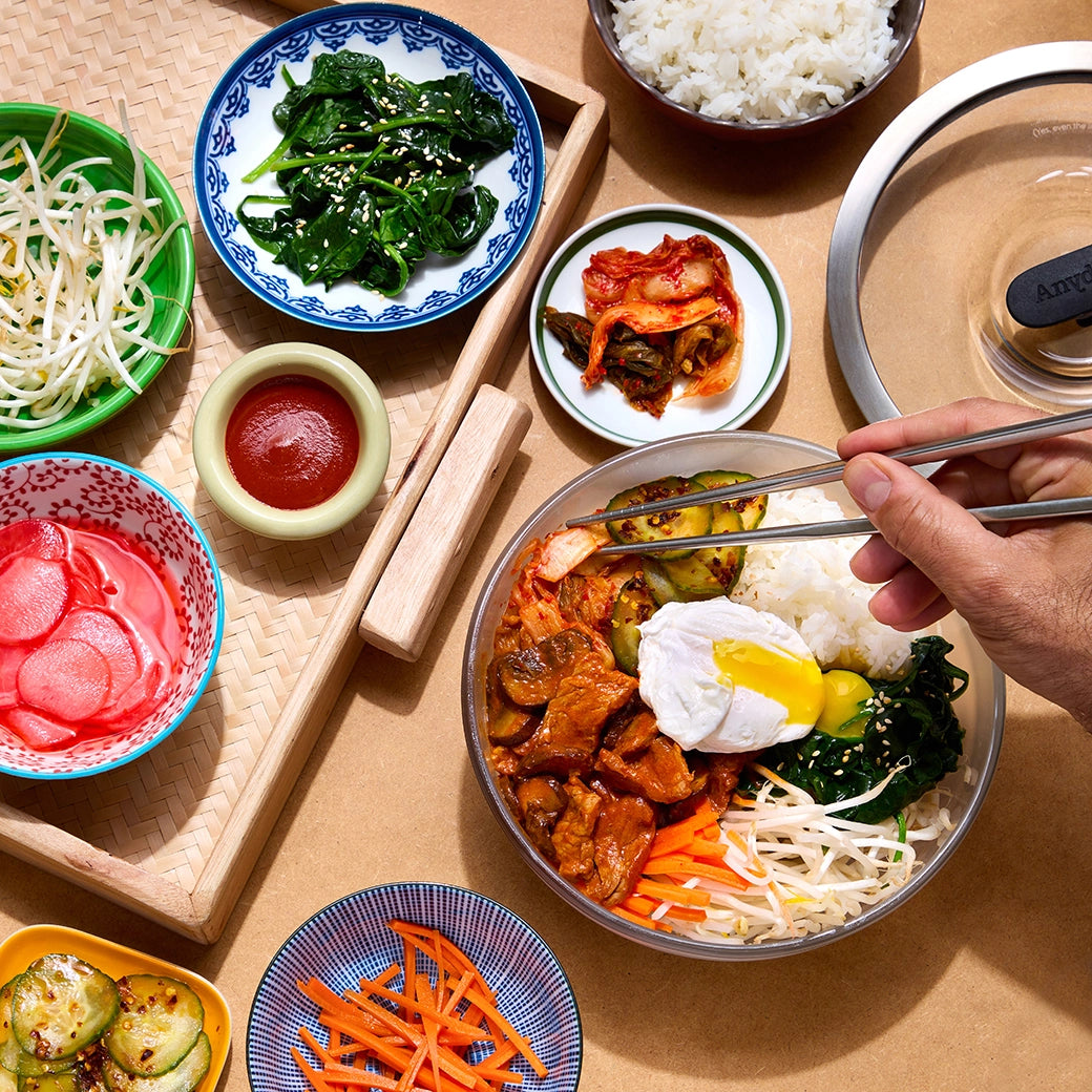 Bibimbap with colorful vegetables, beef, and a poached egg, served in a clear glass Anyday dish, surrounded by various side dishes and sauces.