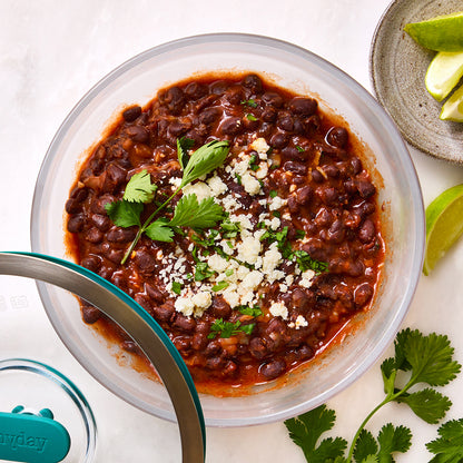 Spicy black bean bake served in a round glass Anyday dish, topped with crumbled cheese and fresh cilantro, with lime wedges and cilantro leaves on the side.