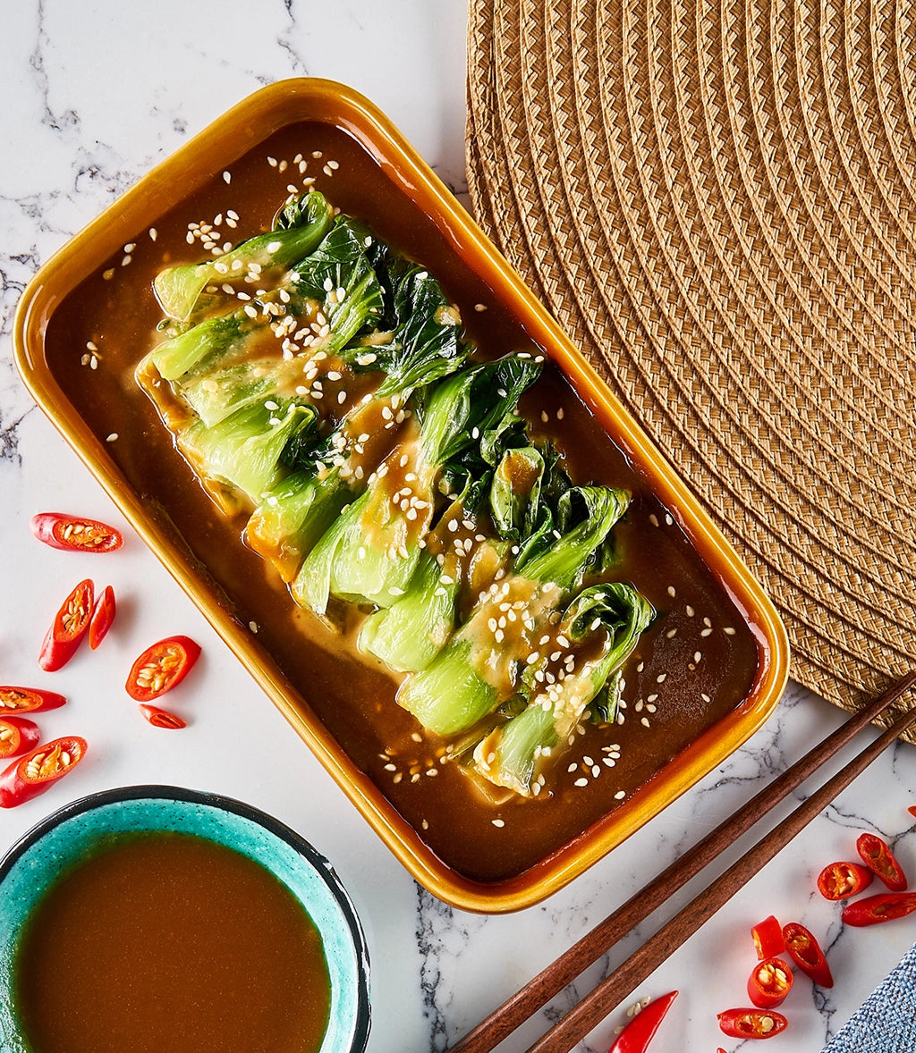 Bok choy cooked in a glass Anyday dish with peanut sauce garnished with sesame seeds, and accompanied by sliced red chili peppers and a bowl of extra sauce.