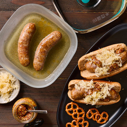 Juicy sausages poached in beer inside an Anyday Square Dish, shown alongside fully assembled sausage sandwiches with sauerkraut and mustard, plus a side of pretzels on a wooden table.
