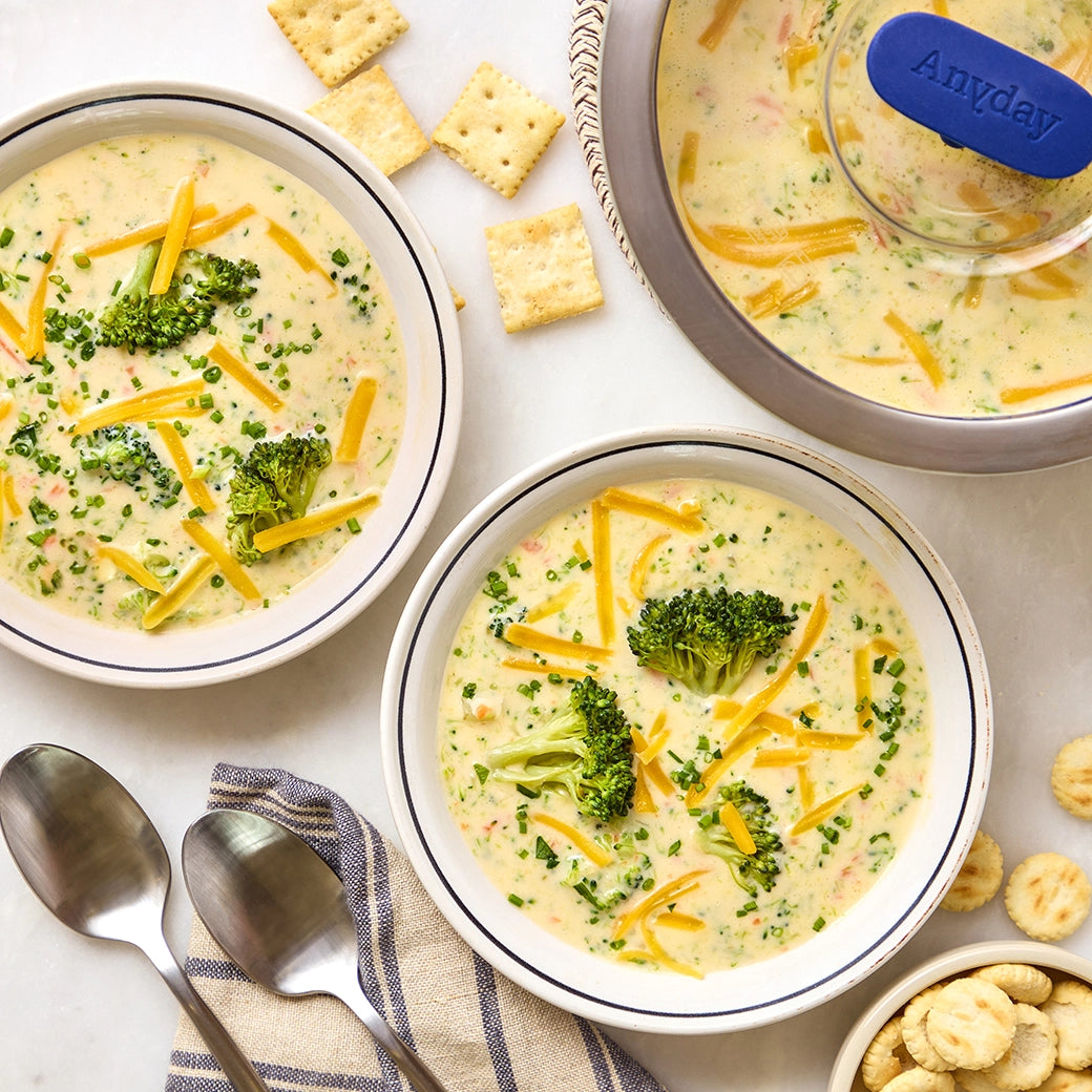 Broccoli cheddar soup made in a glass Anyday dish topped with shredded cheese and broccoli florets, served in bowls, with crackers, and spoons on the side.