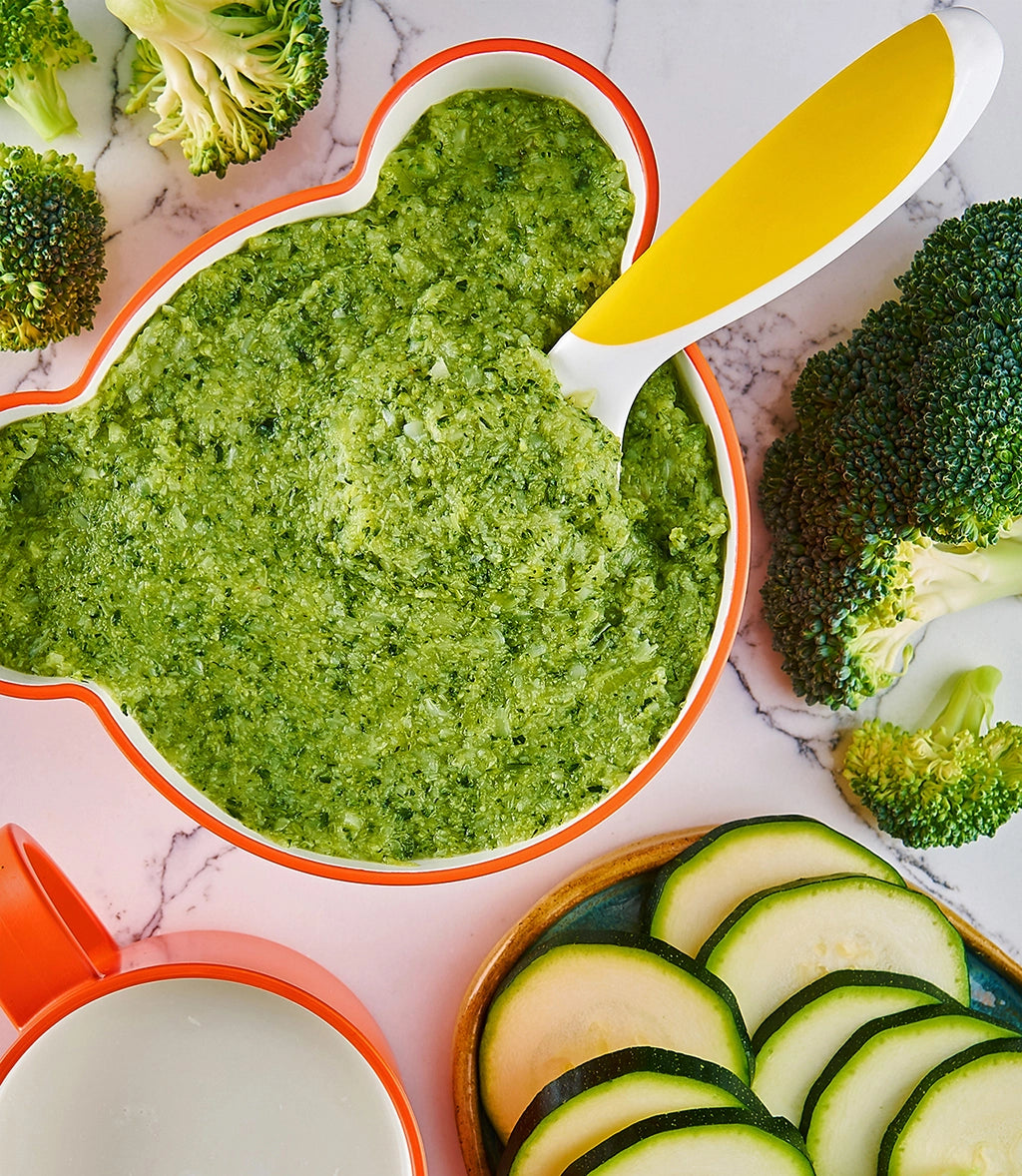 Broccoli and zucchini puree made in a clear glass Anyday dish, accompanied by a blue spoon and fresh broccoli florets on the side.