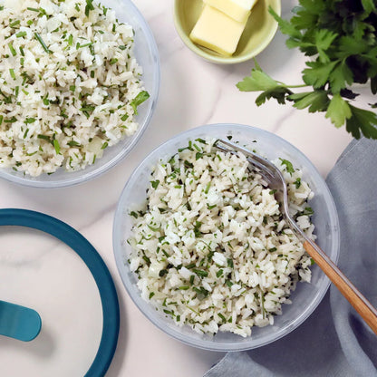 Cooked white rice mixed with fresh chopped herbs served in a clear Anyday dish, with a fork resting inside and a lid and butter nearby on a light countertop.