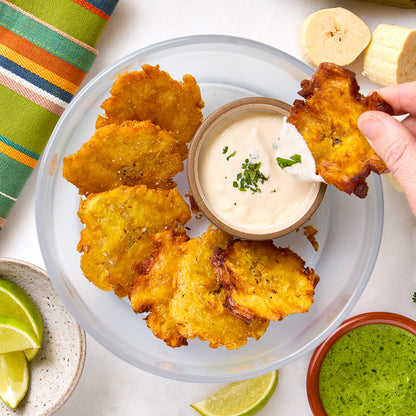 Golden tostones arranged in an Anyday dish with a small bowl of garlic dipping sauce in the center. 