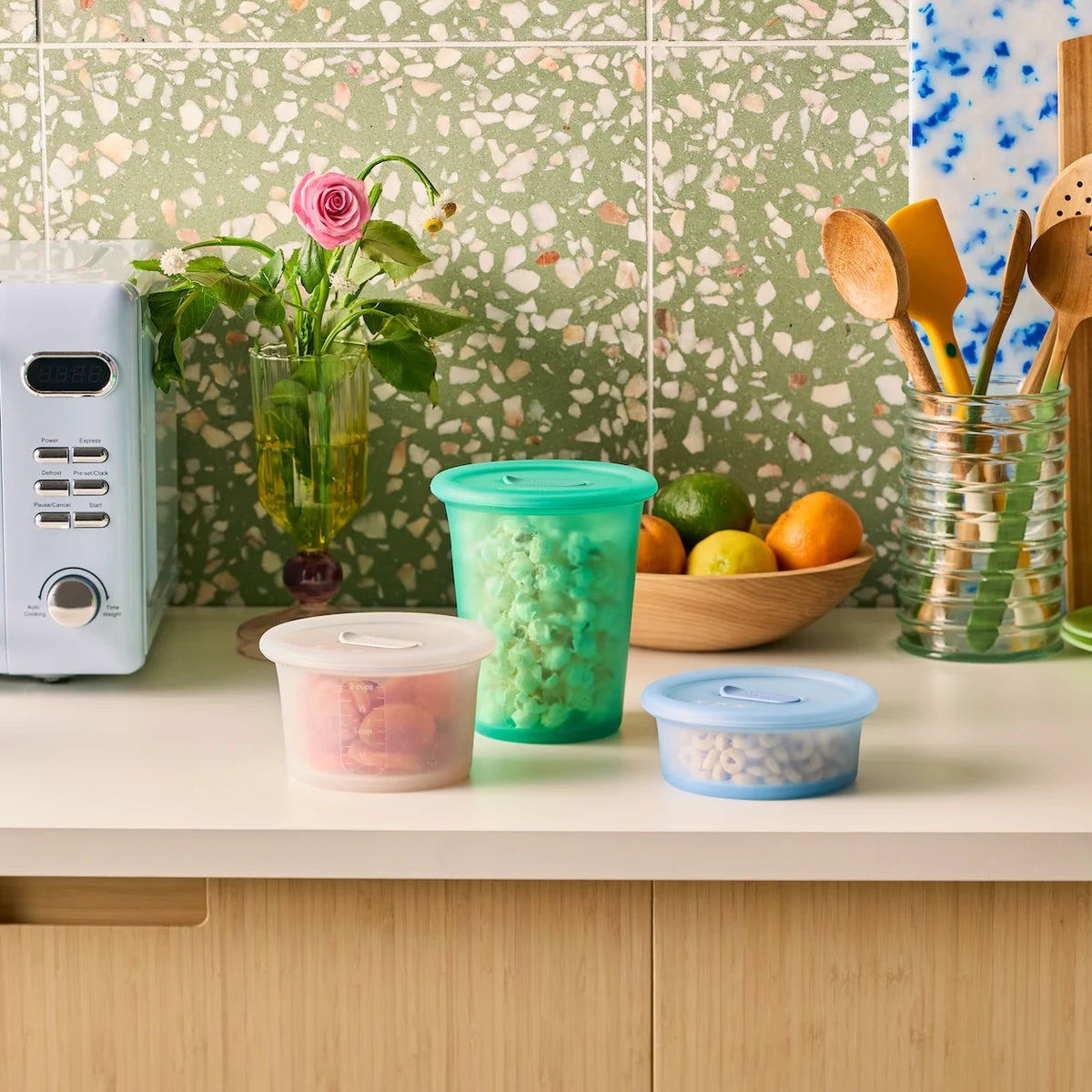 An Anyday silicone Anydeli Bundle set of three food storage canisters on a kitchen counter: a tall green container of popcorn, a small blue container of cereal, and a translucent container of dried fruit.